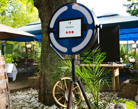 Akku-betriebene Fotobox für Outdoor-Firmenevents Eine akku-betriebene Fotobox steht draußen unter einem Baum in einem Garten, der für eine Veranstaltung dekoriert wurde. Im Hintergrund sind Sonnenschirme und Tische zu sehen.