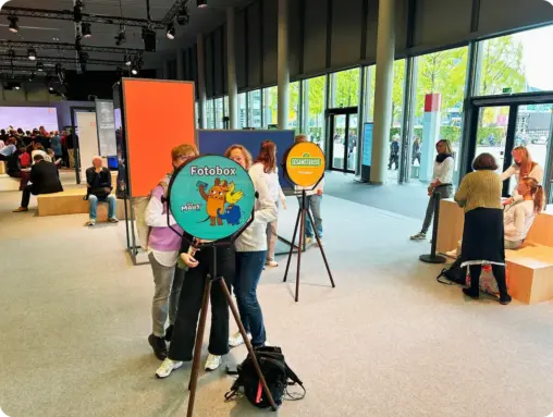 Zwei Mitarbeiterinnen stehen lächelnd hinter einer Fotobox mit "Die Maus"-Branding in einer hellen Messehalle; im Hintergrund ein weiterer Aufsteller der Sesamstraße.