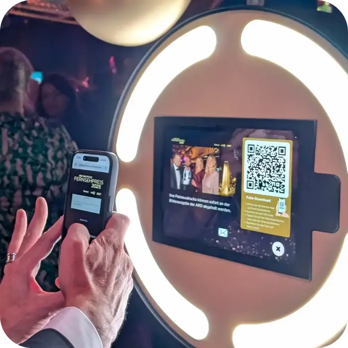Digitaler Bild-Download via QR-Code Nahaufnahme von Händen, die ein Smartphone halten. Der Gast scannt gerade einen QR-Code vom Display der Fotobox ab. Auf dem Handydisplay ist bereits die gebrandete Landingpage 'Der Deutsche Fernsehpreis 2025' mit Login-Maske zu sehen.