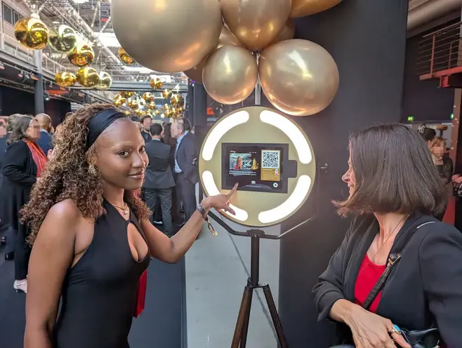 Gäste bedienen die AIR CORE Fotobox Zwei elegant gekleidete Gäste bedienen lachend den Touchscreen der AIR CORE Fotobox auf der Aftershow-Party. Im Hintergrund ist eine festliche Dekoration aus goldenen Luftballons zu sehen.