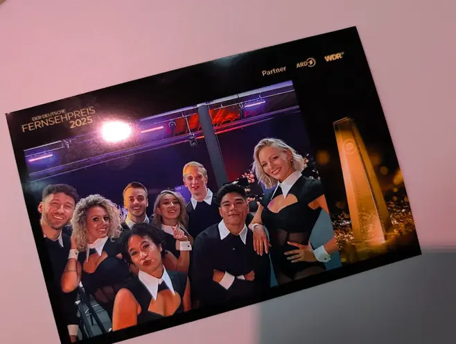 Fotoausdruck Layout Deutscher Fernsehpreis Nahaufnahme eines hochglänzenden Fotoausdrucks. Das Bild zeigt eine Gruppe von Tänzern und Performern. Das Layout beinhaltet das goldene Logo des Deutschen Fernsehpreises 2025, die Trophäe sowie die Partner-Logos von ARD und WDR.